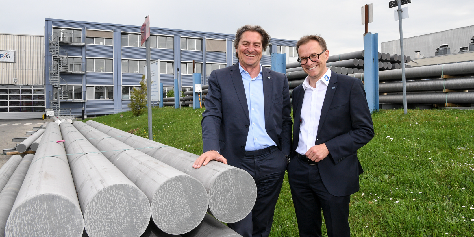 Zwei Personen vor dem Verwaltungsgebäude neben Aluminiumbarren und in der Wiese