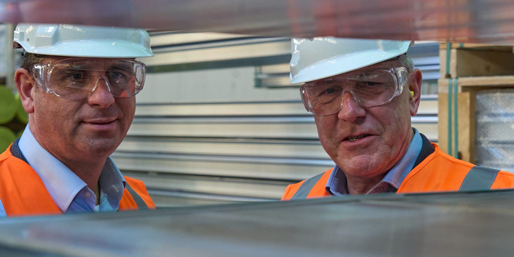 Zwei Männer in orangener Warnweste mit Schutzbrille und Helm prüfen Aluminiumprofile