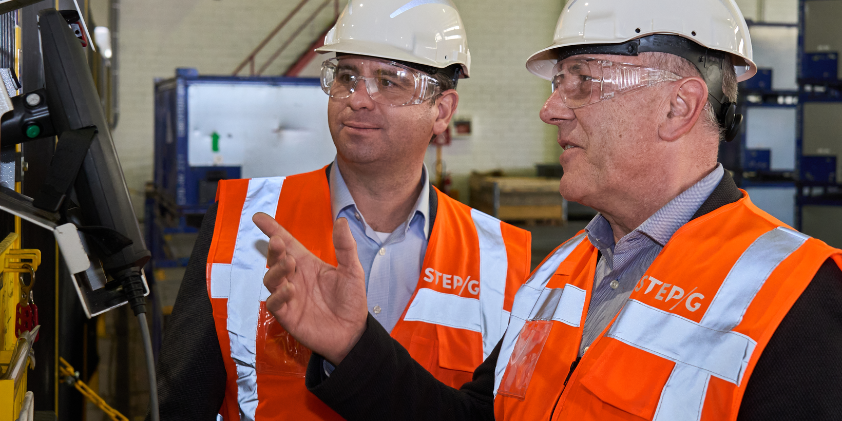Zwei Männer in orangener Warnweste mit Schutzbrille und Helm vor einem Bediendisplay in der Produktion