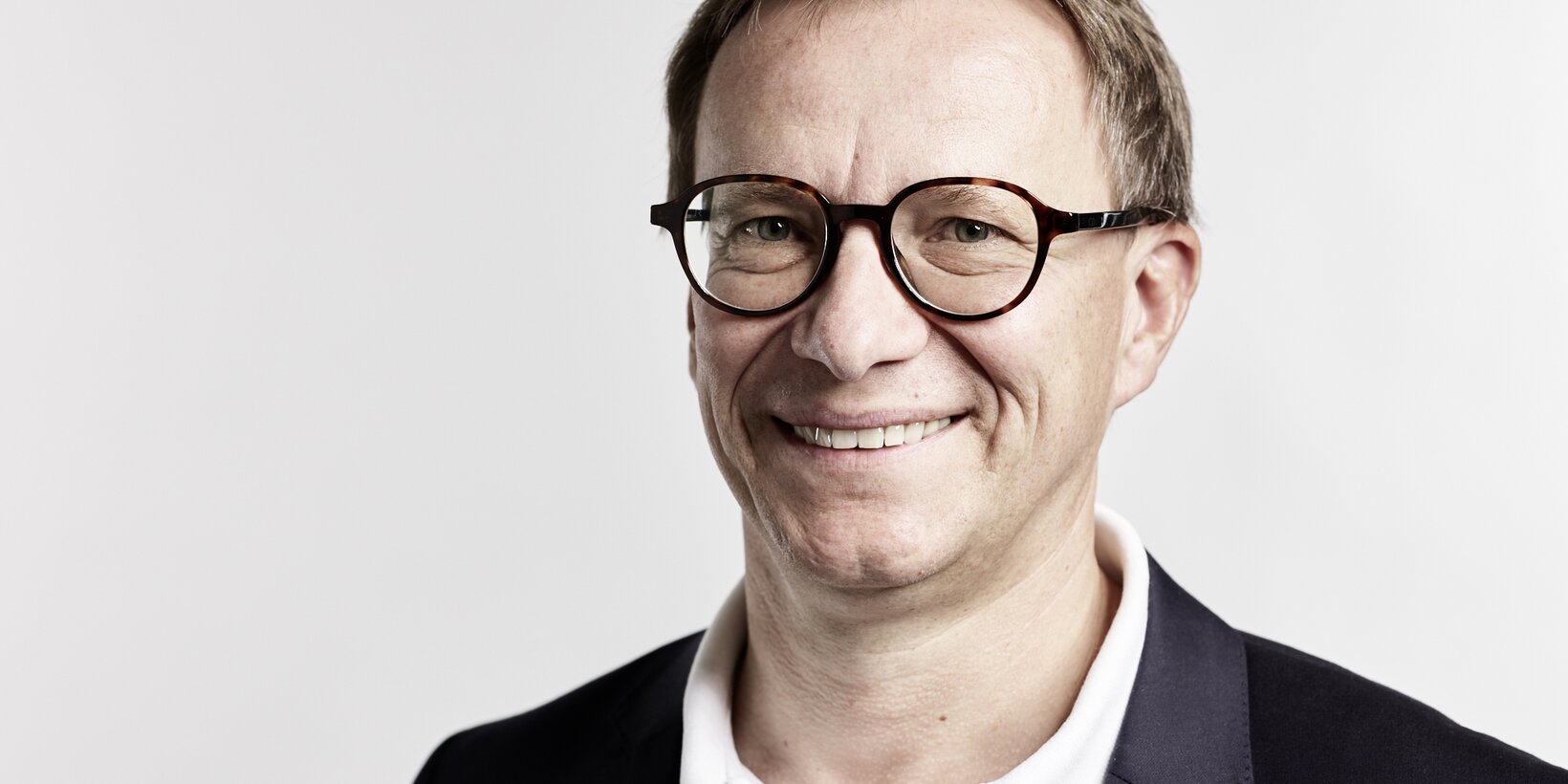 Portraitbild einer männlichen Person im schwarzen Anzug mit weißem Hemd vor hellgrauem Hintergrund