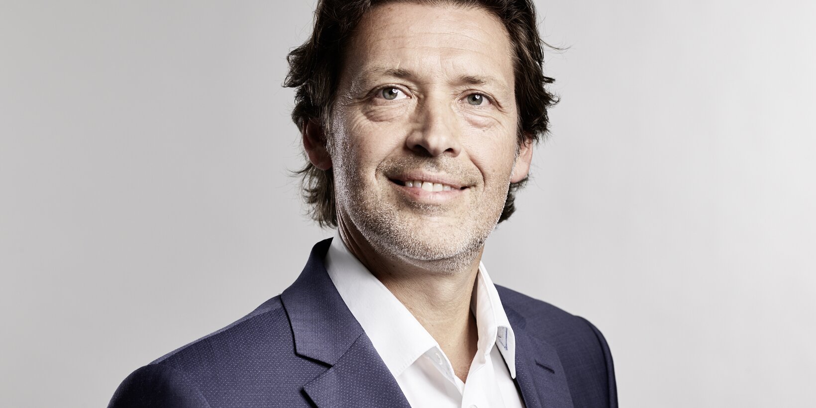 Portraitbild einer männlichen Person im blauen Anzug mit weißem Hemd vor hellgrauem Hintergrund