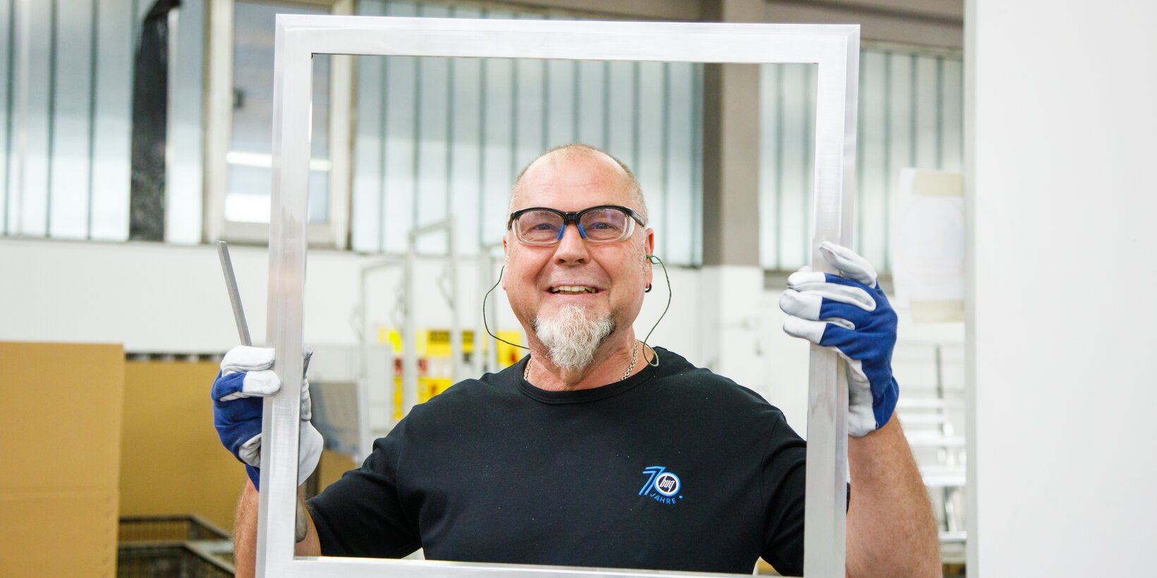 Mitarbeiter aus der Rahmenfertigung von BUG hat eine Aluminiumrahmenkonstruktion in der Hand wie einen Bilderrahmen und lacht